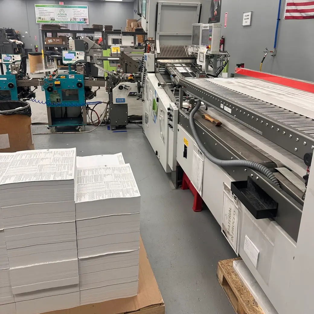 Stacks of printed sheets on a pallet beside the folding line, with the ADIYA PHARMA banner on the far wall